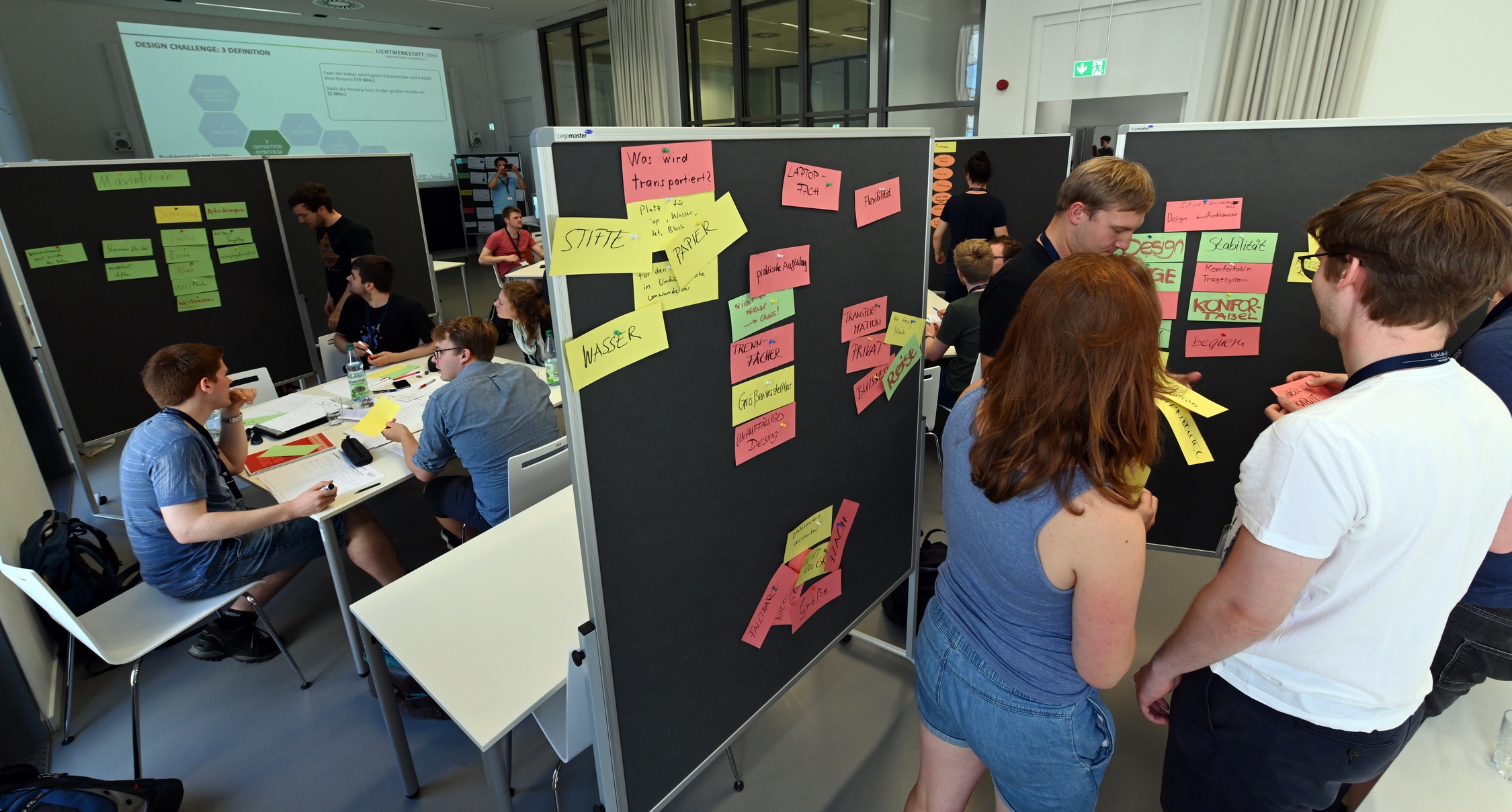 Gruppen von jungen Leuten stehen vor Pinnwänden mit bunten, beschrifteten Zetteln und diskutieren