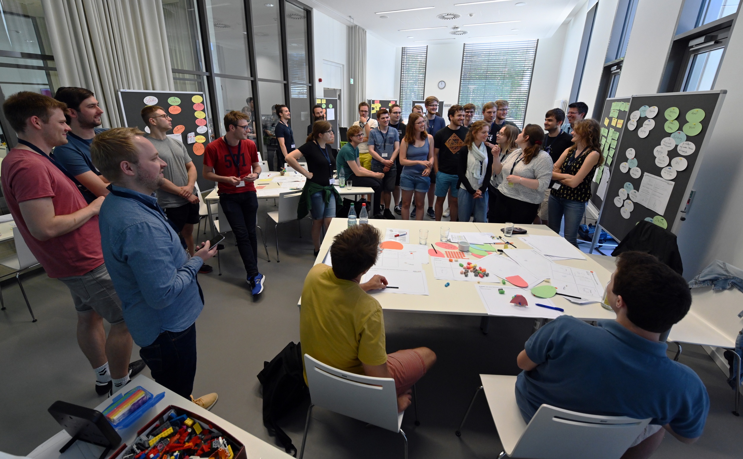 Viele junge Leute stehen in einem Raum mit Pinnwänden, an denen bunte Zettel heften, und diskutieren.
