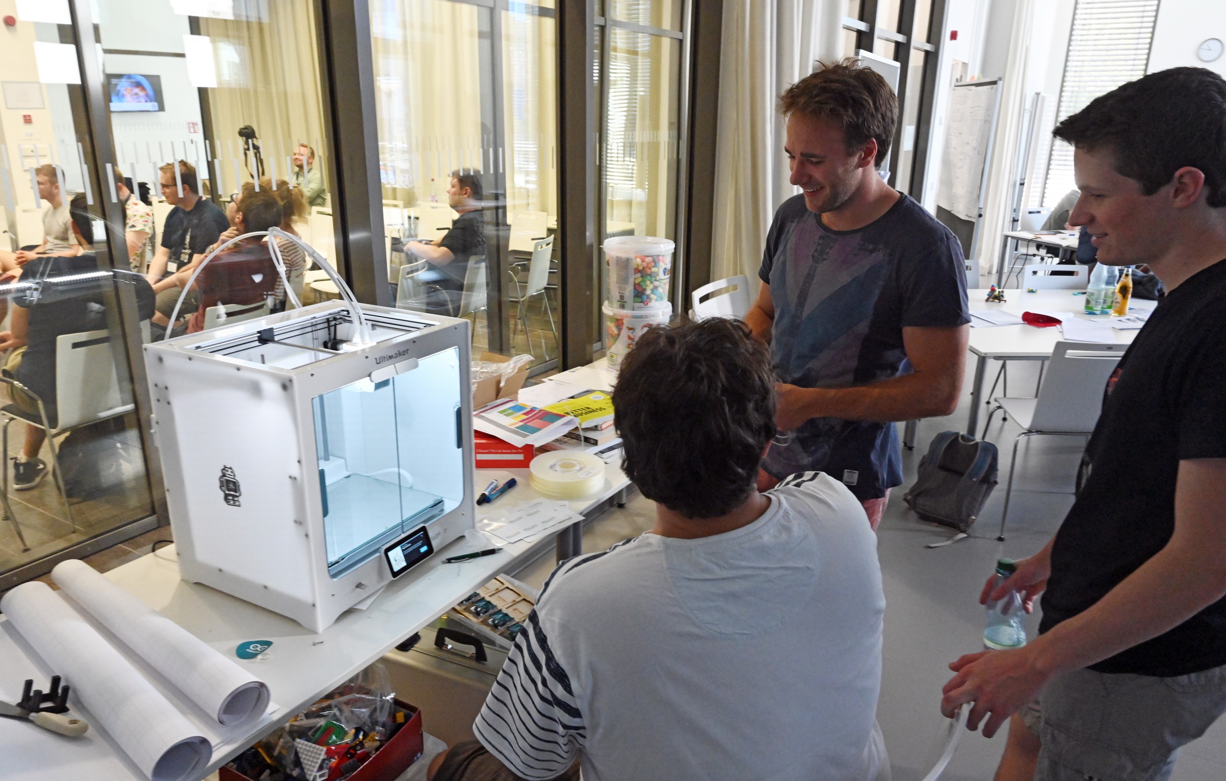 Drei junge Männer stehen vor einem Tisch mit einem 3D-Drucker, einem quadratischen Kasten mit Öffnung oben und einer Glasscheibe an der Frontseite