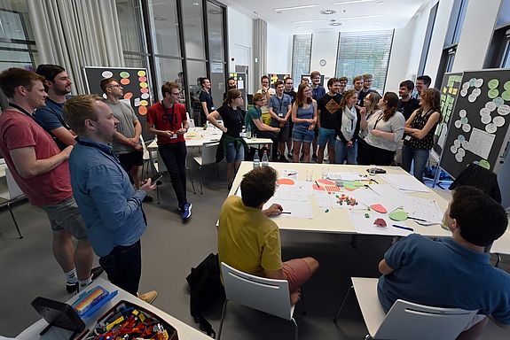 Viele junge Leute stehen in einem Raum mit Pinnwänden, an denen bunte Zettel heften, und diskutieren.