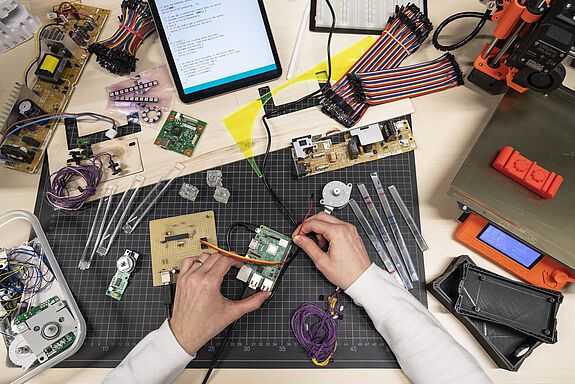 zwei Hände, die eine Platine mit Kabeln und einer Steckplatte verbinden, auf dem Tisch liegen technische Utensilien