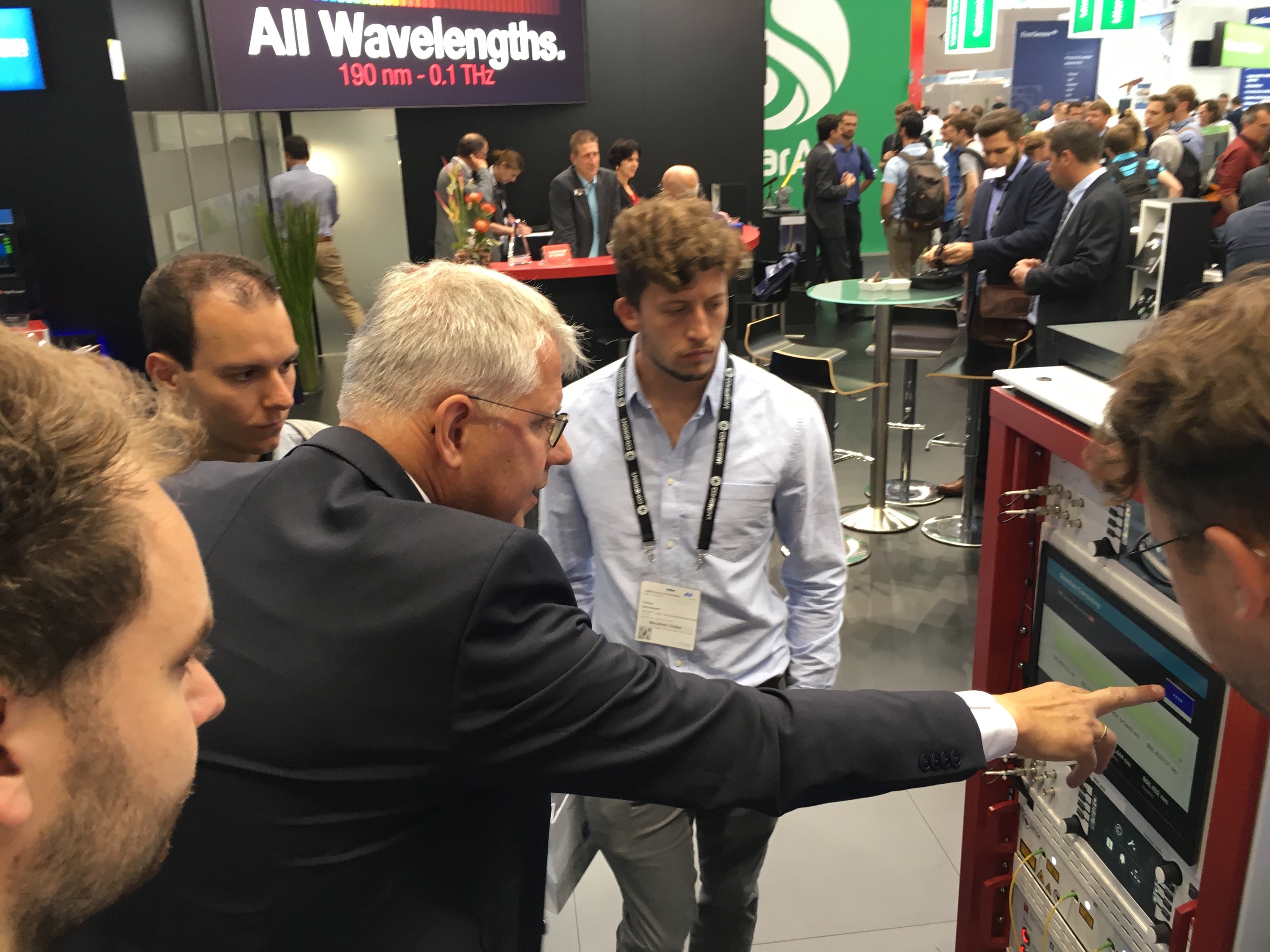 Gruppe von jungen Menschen auf einem Messestand, ein Herr im Anzug deutet auf ein Display und erklärt ihnen etwas